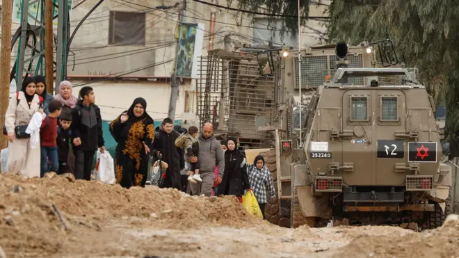 فلسطينيون يسيرون بجوار مدرعات إسرائيلية أثناء مغادرتهم مخيم جنين في الضفة الغربية التي تحتلها إسرائيل، في 22 يناير/كانون الثاني 2025