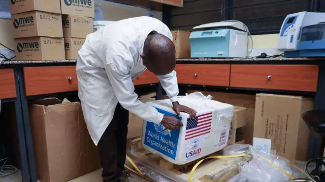 Umugabo wunamye utaboneka mu maso, afashe ku isanduku yanditseho amagambo 'USAID' na 'World Health Organization'. Yambaye itaburiye y'umweru, inyuma ye hari ibikarito. 