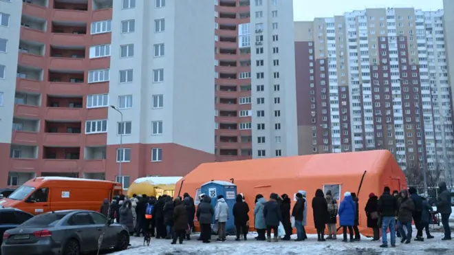 More than a dozen people in winter clothing queue up outside a tent. There is ice on the ground, a few vehicles are parked on the left and tall residential buildings are in the background