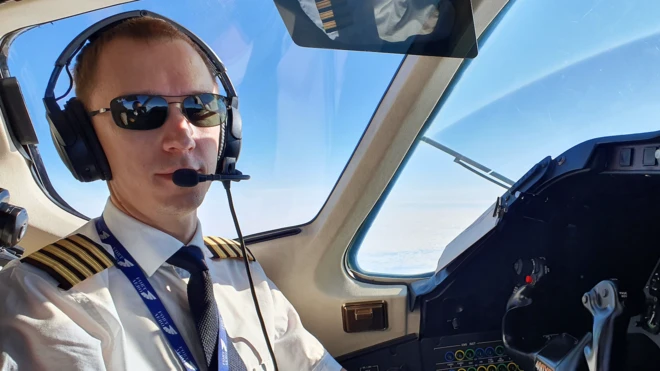 Artur Rodionov sentado na cabine de comando de uma aeronave, aparentemente em voo, tirando uma selfie. Ela usa uniforme de piloto com camisa branca, gravata escura e ombreiras com listras douradas, além de óculos escuros e um headset com microfone. Pela janela da cabine é possível ver o céu azul e uma camada de nuvens abaixo da aeronave. Parte do painel de instrumentos e dos controles do cockpit aparece à direita da foto, reforçando o ambiente de pilotagem profissional.