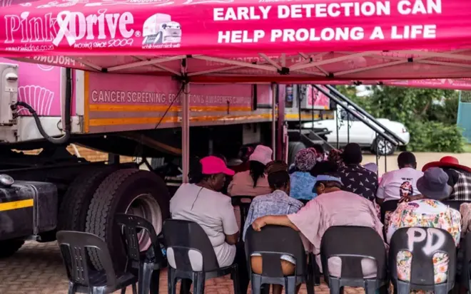 تظهر الصورة مجموعة من الأشخاص المجتمعين، ويافطات تحمل شعارات تدعم الكشف المبكر عن السرطان.