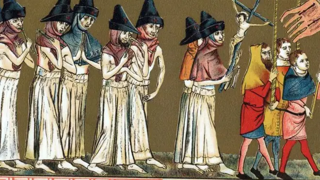 Beyaz cübbeli ve sivri şapkalı sekiz kişi, renkli kıyafetler giymiş üç adamın arkasında çıplak ayakla bir geçit töreninde yürüyor. Adamlar bayrak taşıyor.
