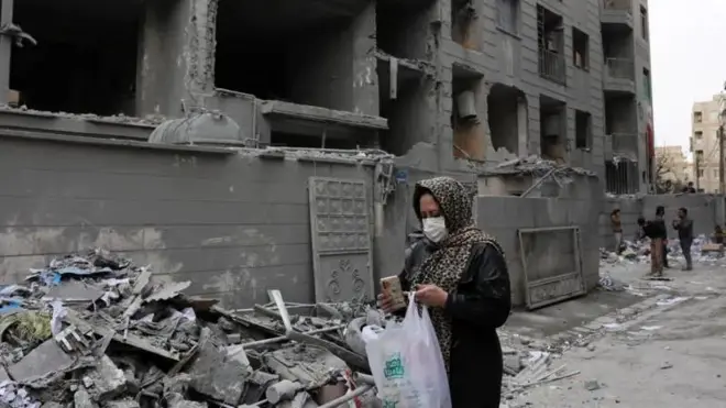 Umugore wambaye agapfukamunwa n'ikanzu y'umukara, witandiye mu mutwe, nyuma y'ibitero bya Israel mu burengerazuba bwa Irani. Mu kiganza kimwe afite telefone igendanwa naho mu kindi kiganza afite ishashi y'umweru n'ubururu irimo ibintu imbere. 