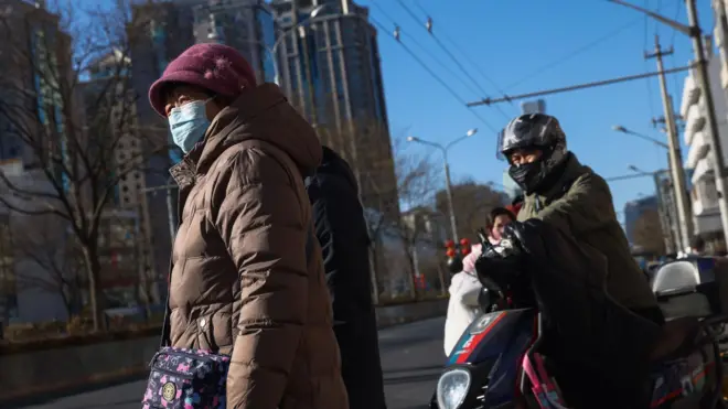 बिजिंगच्या रस्त्यावर मास्क घालून उभी असलेली एक वृद्ध महिला