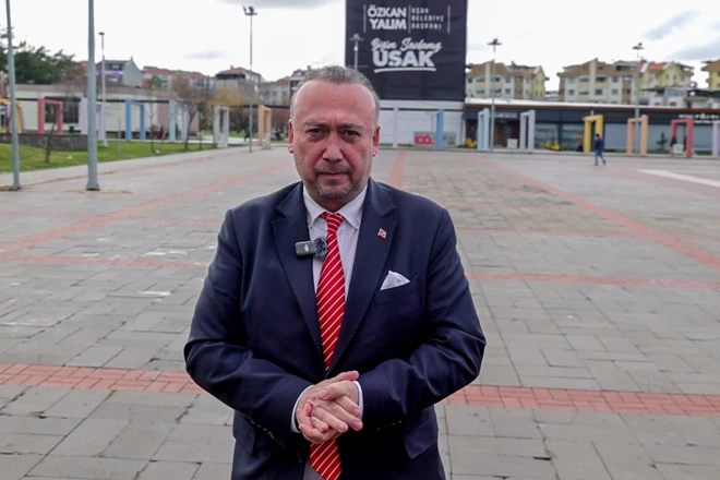 Özkan Yalım kent meydanında ayakta duruyor, ellerini birbirine bağlamış, kameraya bakıyor. 
