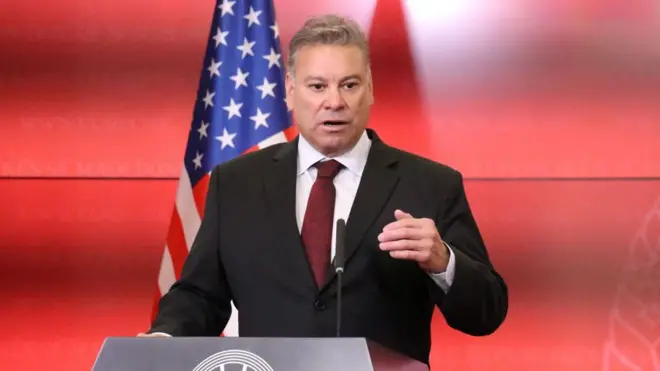 Gabriel Escobar  discursando com bandeira americana ao fundo