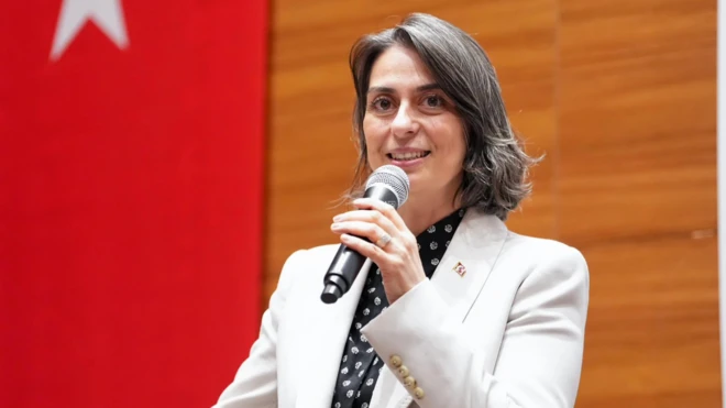 Sinem Dedetaş elinde mikrofonla sahnede konuşuyor. Beyaz bir takım giymiş, arkasında Türk bayrağı var.