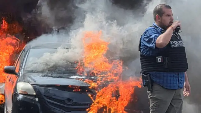 Un vehículo incendiado durante los disturbios por la muerte de "El Mencho", a su lado, un policía de civil con un chaleco antibalas y una pistola al cinto a habla por radioteléfono