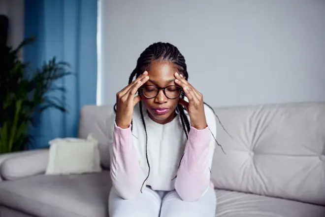 Aworan obinrin kan to joko pẹlu awo oju loju rẹ, o fi ọwọ mejeeji di ori mu