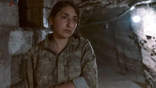 صورة بورتريه لآريين داخل نفق. ترتدي زياً عسكرياً مع عقد وسوار، وتضع ذراعاً مطوية عند خصرها، بينما تنظر إلى ما وراء الكاميرا بنظرة حالمة. ويضيء مصباح مثبت على جدار النفق المكان من خلفها.