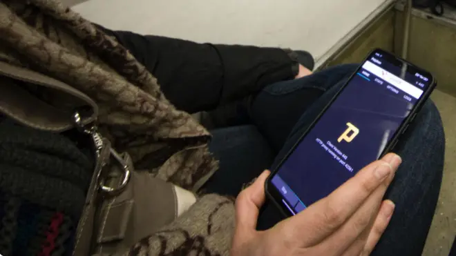 دست مردی که در اتبوس نشسته گوشی تلفن را که روی زانویش قرار دارد گرفته است. روی گوشی اپ سایفون باز است.