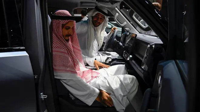 在一场车展上，两名身穿长袍、头戴头巾的男子，分别坐在一辆中国制造的汽车的驾驶座位和前排乘客座位。