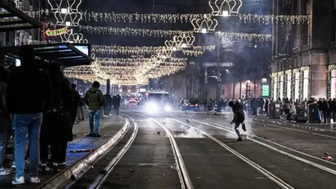 Okićene ulice, ljudi na trotoarima, dim od vatrometa po šinama 