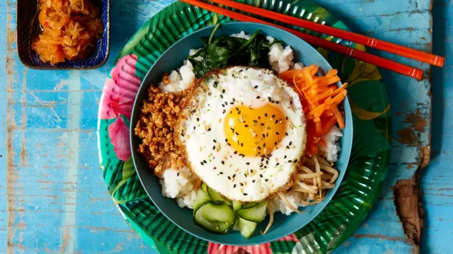 Bibimbap, a Korean dish featuring rice and various vegetables such as carrots, spinach, bean sprouts and a fried egg on top, served with chopsticks, and a side dish of kimchi.
