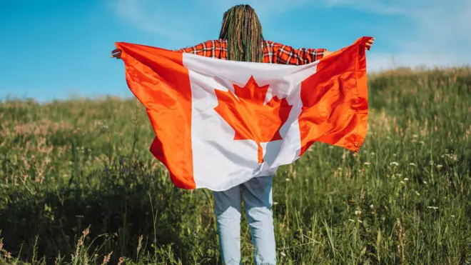 A young girl backing di camera as she hold Canada flag while she stand for inside a garden wit flower