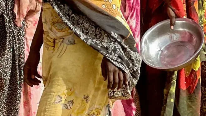 Image recadrée de plusieurs femmes du Darfour vêtues de robes colorées, sans montrer leur visage. L'une d'elles tient un plat en argent vide dans sa main. La photo a été prise en avril 2025.