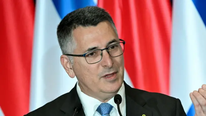 Gideon Saar stand for front front of a row of flags and speak into microphone