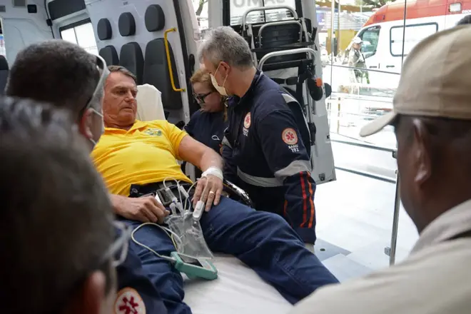 Fotografia colorida mostra o ex-presidente Jair Bolsonaro deitado na maca dentro de ambulância em Natal, com paramédicos ao seu redor. Ele veste uma camisa amarela do Brasil