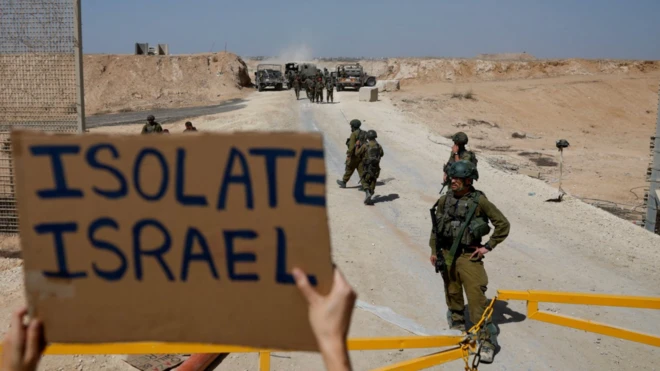Bir gösterici İsrail askerlerinin göründüğü bir karede onlara karşı önünde bir pankart tutuyor. Sadece askerleri ve pankartı görüyoruz