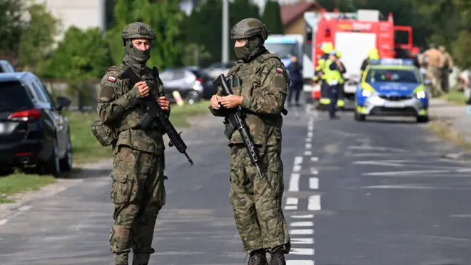 Dos integrantes del ejército polaco, vestidos con ropa de militar y armas largas están en primer plano en medio de una carretera, ataviados con cascos y pasamontañas que les tapa la mitad de la cara. Al fondo están los servicios de emergencia, un coche de policía, un camión de bomberos y, muy a lo lejos, se distinguen figuras humanas. 