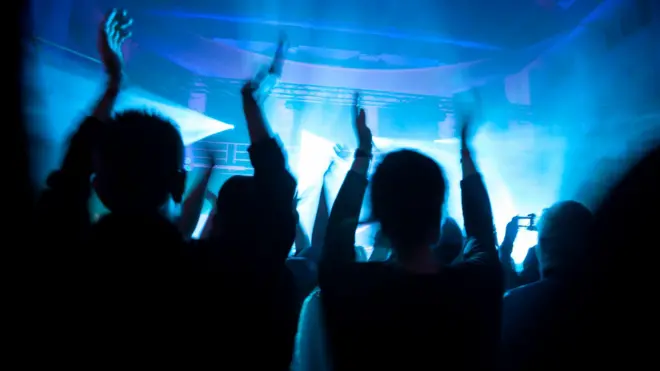 Cилуети людей у натовпі, які підняли руки й аплодують. Перед ними яскраве синє сценічне світло, що розмиває контури та створює ефект енергійного концертного або клубного виступу. Атмосфера динамічна й наповнена рухом.