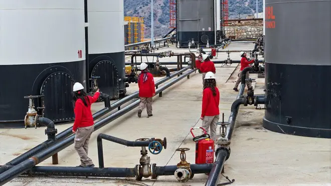Kadın operatörler petrol dağıtım tesisinde boruların etrafında çalışıyor hepsi kırmızı üst, krem rengi kargo pantolon giyinmiş ve baretliler. 