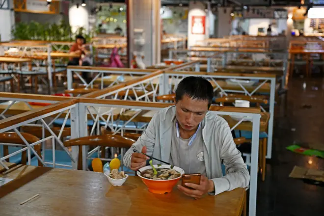 Man with a bowl of food sitting in a canteen looking at his phone