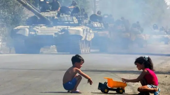 جاده خاکی با ردیف تانکها و دو کودک در کنار جاده در حال بازی با یک کامیون اسباببازی