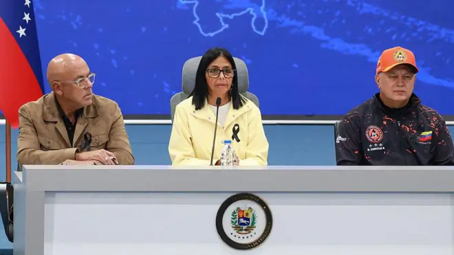 Jorge Rodríguez, Delcy Rodríguez y Diosdado Cabello sentados en una mesa con un micrógono en el centro, fondo azul con la silueta del mapa de Venezuela y una bandera de Venezuela a la izquierda.
