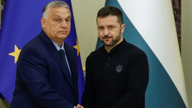 Виктор Орбан и Владимир Зеленский пожимают друг другу руку, глядя в камеру с кислыми лицами