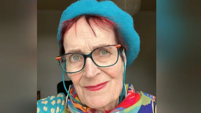 A selfie of Mary Thornton. She has short red hair and blue eyes. She is wearing a bright blue beret and a colourful shawl. She also wears glasses with a blue and orange frame. She is slightly smiling.