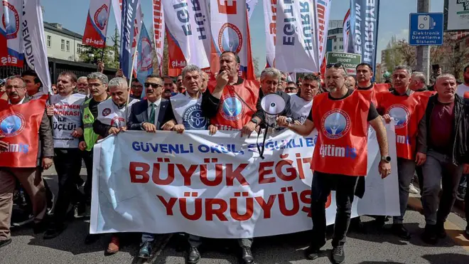 öğretmenler "güvenli okul, sağlıklı eğitim, büyük eğitim yürüyüşü" yazılı pankartlarla yürüyor, beyaz ve kırmızı yelekler giymişler, önde bir erkek elinde megafonla konuşuyor, arkalarında da sloganlı beyaz ve mavi bayraklar var