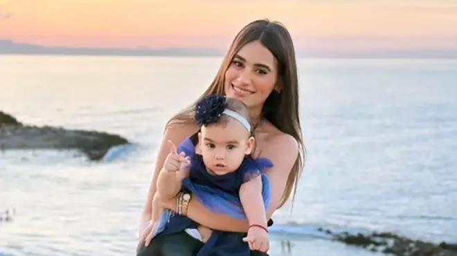 Una mujer de cabello largo y oscuro sonríe mientras sostiene a una niña pequeña vestida con un vestido en la playa, con el atardecer de fondo.