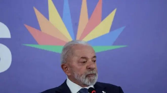 Foto de Lula perto de um microfone diante de um fundo azul e de uma figura multicolorida com formas geométricas parecendo uma flor