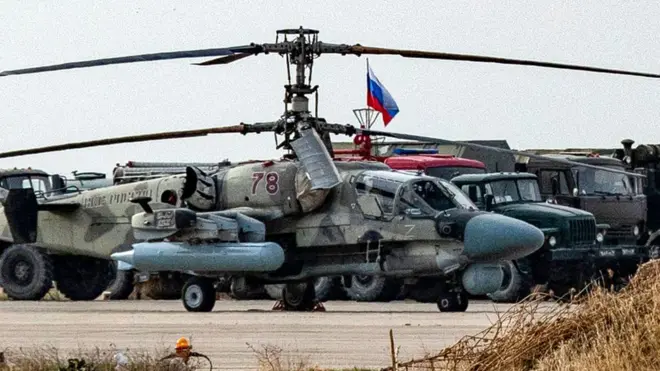 Askeri helikopter yanında iki askeri kamyon havaalanında park edilmiş, aralarında bir Rus bayrağı görünüyor. 