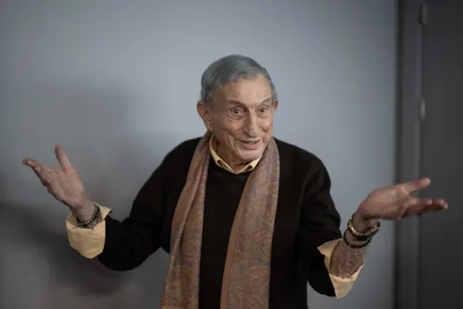 Haldun Dormen, siyah kazak ve kahverengi, desenli bir atkı giymiş halde ellerini iki yana açıp, gülümserken 