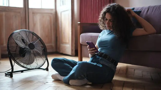 Młoda kobieta patrzy w ekran telefonu. Siedzi na drewnianej podłodze, opierając łokieć o siedzenie kanapy. Obok niej stoi okrągły czarny wentylator.