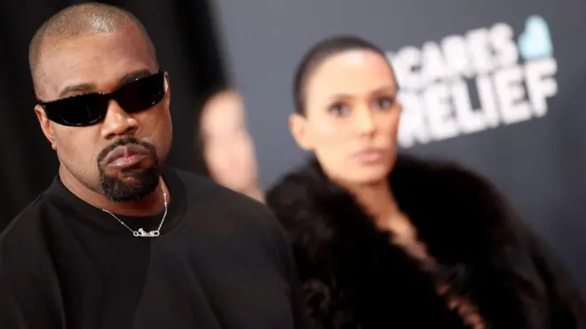 Kanye West areba muri 'camera', inyuma ye ku ruhande hari umugore we Bianca Censori, na we areba muri 'camera'. 
