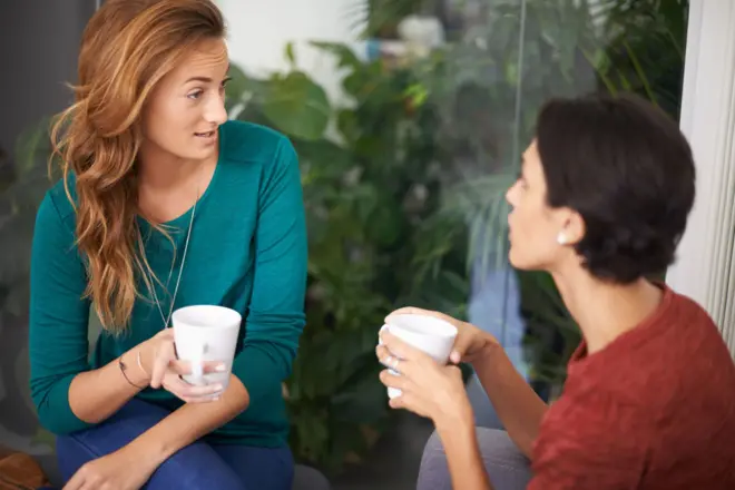 Duas mulheres brancas conversam segurando canecas nas mãos, uma delas faz carabet bola apostaincredulidade