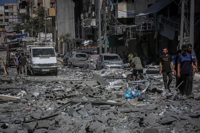 A street in the Rimal neighbourhood of Gaza City is littered with rubble. Many destroyed cars can be seen lining the streets as peole congregate in small groups