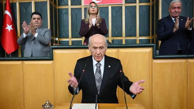 Bahçeli ellerini kürsüde iki yana açmış durumda, arkasında üç kişi onu alkışlıyor