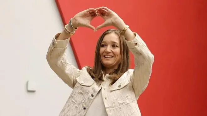 Beyaz kıyafetli Durbay elleriyle kalp işareti yapıyor.