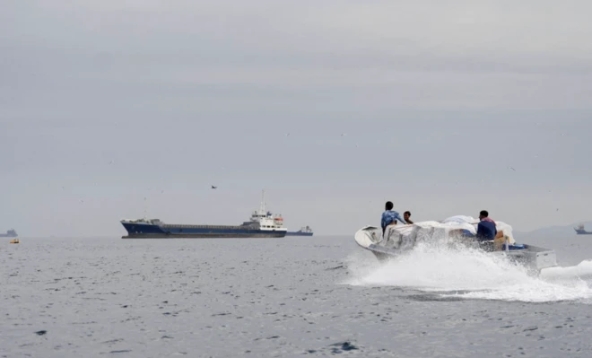 Üç kişinin içinde olduğu bir deniz motoru hızla ilerliyor. Arkada büyük bir tanker var.