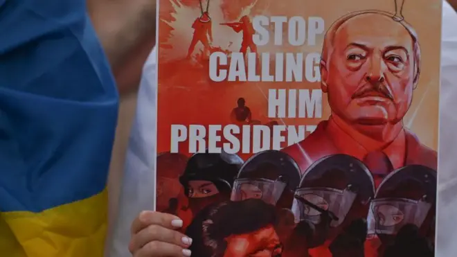 Справа женская рука держит рисунок, на котором Лукашенко и спецназ на фоне надписи по-английски "Прекратите называть его президентом", а слева кто-то с флагом Украины