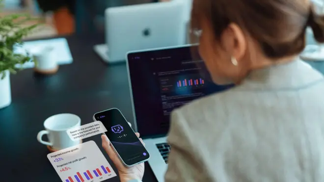 Over the shoulder image of a businesswoman wearing a suit. She is looking at an AI chatbot on her smartphone.