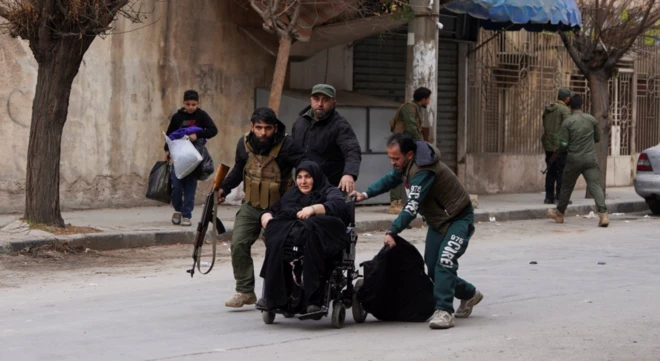 Tekerlekli sandalyedeki bir kadın ona yardımcı üç kişi tarafından bir araç yolunda itiliyor. Adamlardan biri kalaşnikof tüfek taşıyor. Arkada başka silahlı kişiler uzaktaki bir noktaya bakıyor.