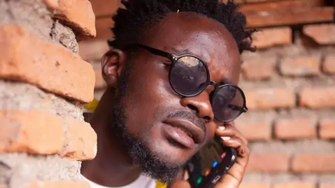 Freddy Mukuza porte des lunettes de soleil rondes et parle au téléphone portable en passant la tête autour d'un mur.