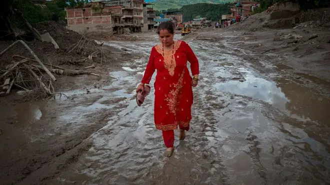 Una mujer camina descalza por un rio de lodo y detrás se ven edificios derruidos.