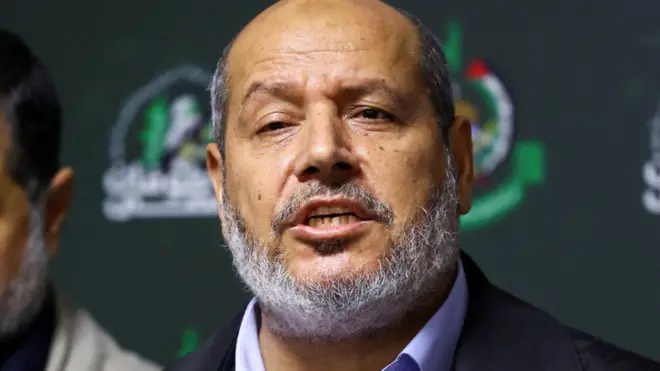 Hamas officials, Khalil al-Hayya, at a news conference podium in Beirut, Lebanon, on 21 November 2023. He is speaking and has a grey beard that runs from his chin to ears and a balding head. Al-Hayya is wearing an open-collared purple shirt and a dark blue casual outdoor jacket.