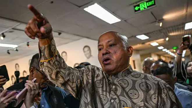 Philippine Senator Ronald Bato Dela Rosa gestures with his right hand, pointing upwards with his index finger while surrounded by journalists in the Senate hall